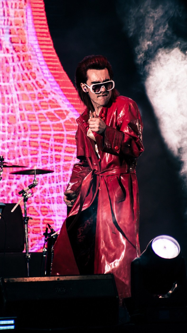 Gaspar Luna en vivo con tapado rojo de charol y gafas blancas sobre el escenario